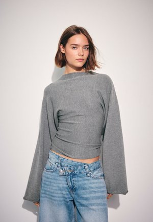 Jeune femme aux cheveux courts bruns portant un haut gris à manches longues et un jean bleu clair ample sur un fond blanc uni.