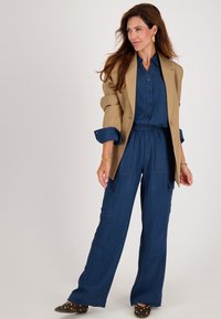 Beige blazer over een blauwe overhemd met knopen en bijpassende wijde broek. De outfit heeft een gestructureerde stof, zakken en gestikte bruine schoenen.