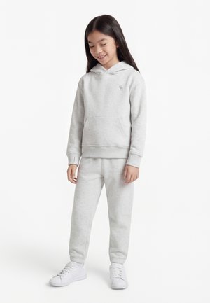 Uśmiechnięta dziewczyna w jasnoszarej bluzie z kapturem i dopasowanych spodniach dresowych oraz białych trampkach, stojąca na tle jednolitego białego tła.