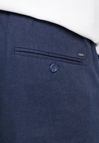 Granatowe lniane spodnie z kieszenią z przodu, zapięciem na guzik i dodatkową metką na pasie. Teksturowany materiał o subtelnej fakturze.