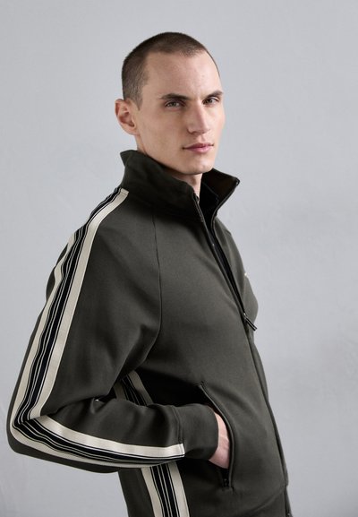 Olivovo zelená bunda na zips s vysokým golierom, s dvoma bielymi a čiernymi pruhmi pozdĺž rukávov a bočných vreciek.