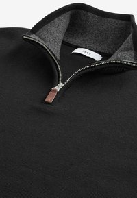 Crna zip-up pulover jakna s visokim ovratnikom. Ima kontrastni sivi unutarnji ovratnik, metalni zatvarač i kožni akcent tag. Teksturirana tkanina.