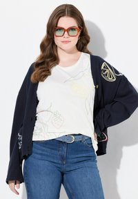 Mørkeblå cardigan med frugt-tema broderi på ærmerne, parret med en hvid skjorte med omrids af frugtdesign, blå bælte og mørke jeans.