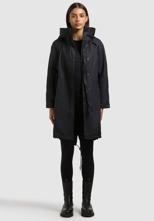 Naine seisab, kandes musta keskmise pikkusega manteliga kõrge kraega, musti retuuse ja musti saapaid, taustal hele ja lihtne taust.