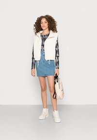 Colete branco acolchoado sobre uma camisa de mangas longas em tie-dye preto e cinza, combinado com uma saia de ganga azul, ténis brancos de cano alto e uma mala clara.