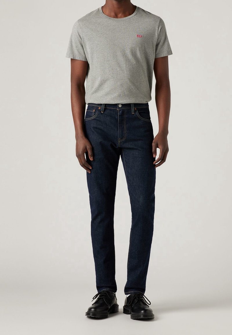 Grå t-shirt med korta ärmar och en liten röd logotyp, kombinerad med mörkblå smala jeans och svarta snörskor. Enkel, avslappnad stil.