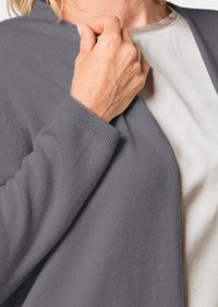 Grijze cardigan van zacht breimateriaal, met lange mouwen, geribde boorden en een ontspannen halslijn over een lichtkleurig shirt.
