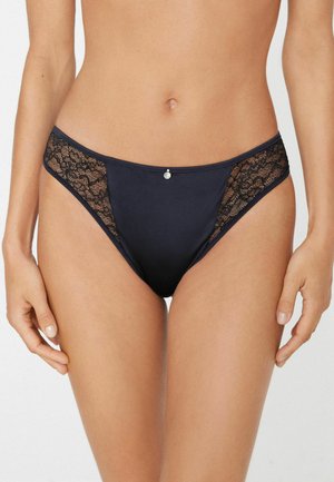 Kobieta nosząca czarne bikini z koronkowymi panelami i małym ozdobnym zawieszką na środku z przodu, pokazana od talii do górnej części ud.