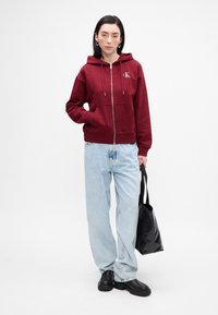 Bordo zip-up dukserica s vezicama, prednjim džepovima, u kombinaciji sa svijetloplave slobodne traperice i crnim platformama, držeći crnu torbu.