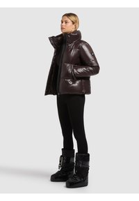 Kvinna klädd i en glänsande mörkbrun vadderad jacka, svarta leggings och svarta vinterkängor som står mot en enkel ljus bakgrund.