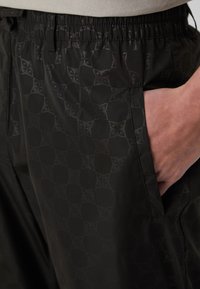 Schwarze Sporthose mit elastischem Bund zeigt ein dezentes geometrisches Muster und Seiten Taschen. Der Stoff wirkt glatt und leicht.