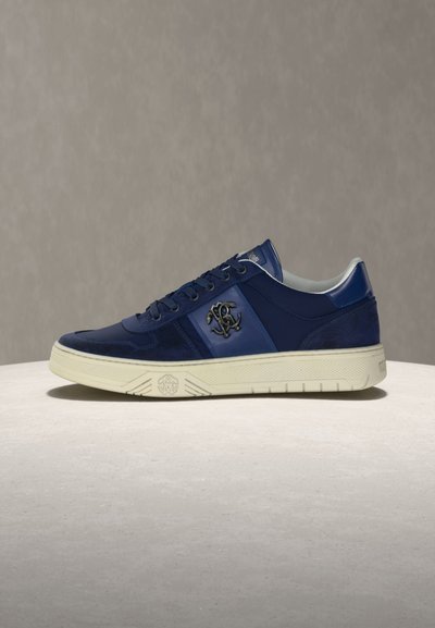 Παπούτσι χαμηλού τύπου σε ναυτικό μπλε με δερμάτινες και σουέντ λεπτομέρειες, σόλα από κρεμ καουτσούκ και μεταλλικό έμβλημα λογότυπου στο πλάι, τοποθετημένο σε ουδέτερη επιφάνεια.