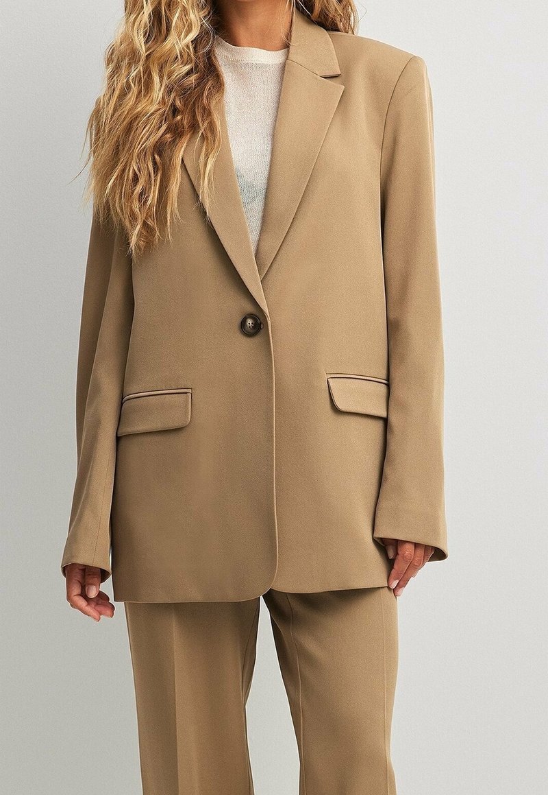 Béžové sako na míru s jedním knoflíkem, s výstřihem do klopy a dvěma předními kapsami. Měkká látka a volný střih. Kalhoty v příslušné barvě.