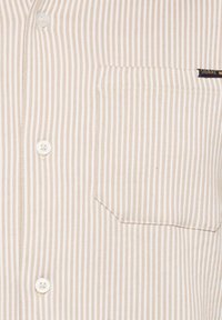 Beige stribet button-down skjorte med en brystlomme, udstyret med hvide knapper og et tekstureret bomuldsstof. Simpelt design med vandrette linjer.