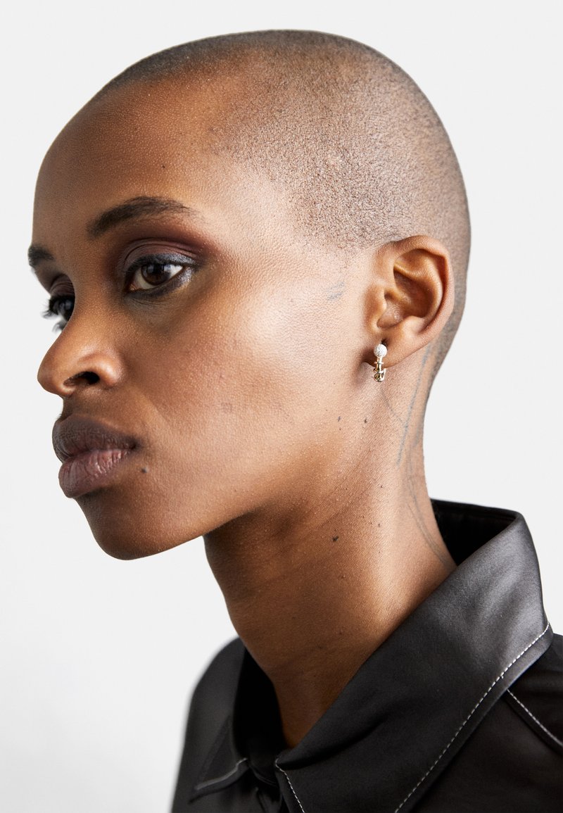 Profile view of a person with a shaved head, wearing a black shirt collar. Features include smooth skin, defined jawline, and a small earring.