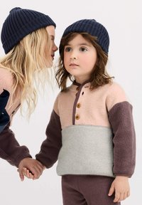 Zwei Kinder halten Händchen; eines flüstert dem anderen zu, während sie passende marineblaue Strickmützen und mehrfarbige Fleece-Oberteile tragen.