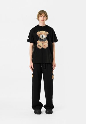 Camiseta negra con un gráfico texturizado de un oso en la parte frontal, combinada con pantalones negros que presentan acentos de oso y detalles de cordón. Calzado casual.