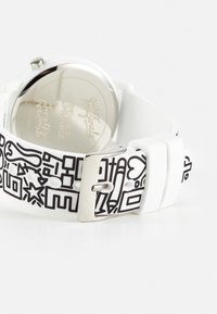 Bracelet de montre blanc avec des griffonnages abstraits noirs et une boucle en argent, montrant le dos de la montre argenté et réfléchissant.