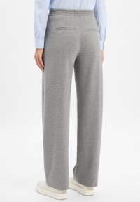 Persoon in losse grijze broek, witte sneakers en een blauw gestreept shirt, staand met de rug naar de kijker op een witte achtergrond.