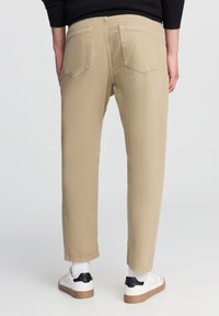 Pantaloni beige a vestibilità rilassata con tasche posteriori, abbinati a sneaker bianche con accenti neri. Il tessuto ha un aspetto testurizzato.