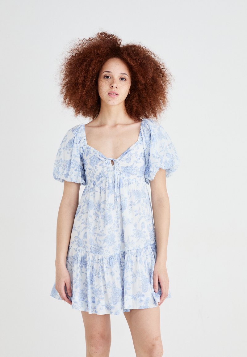 Vestido floral azul com mangas bufantes, decote em V e detalhe em fenda. Apresenta uma bainha em camadas com babados e textura de tecido leve.