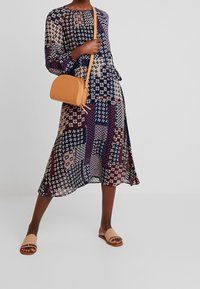 Femme portant une robe midi à motifs patchwork, tenant un sac bandoulière marron clair, portant des sandales beiges, debout jambes écartées sur un sol gris.