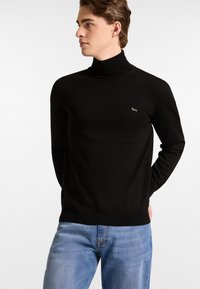 Schwarzer Rollkragenpullover aus glattem Stoff, mit schlankem Schnitt und einem kleinen weißen Logoakzent auf der Brust, kombiniert mit blauen Jeans.
