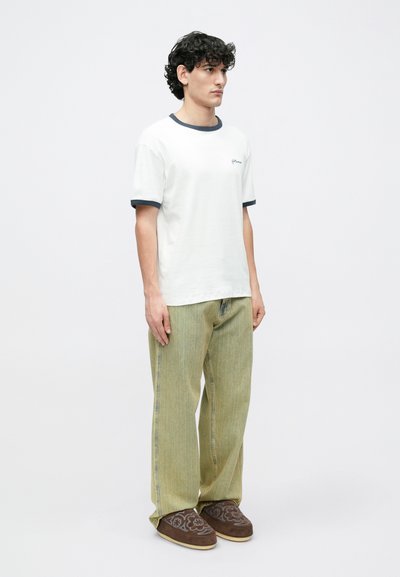 Fehér, rövid ujjú póló tengerészkék gallérdísszel, világoszöld, bőszárú nadrággal és barna, mintás, felhúzható cipővel párosítva.