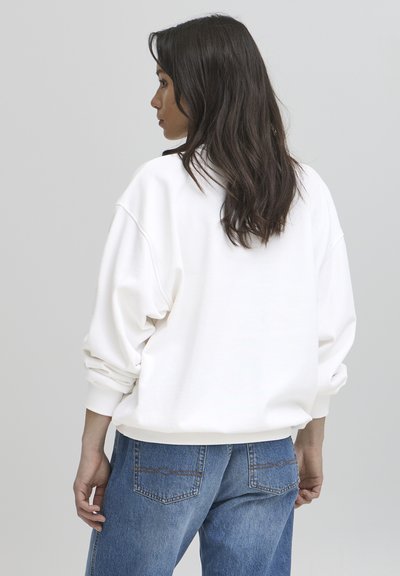 Sudadera blanca de gran tamaño con hombros caídos y puños acanalados, combinada con jeans azules de talle alto que presentan costuras en los bolsillos traseros.