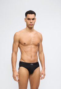 Μυώδης άνδρας με κοντά σγουρά μαλλιά, όρθιος, φοράει μαύρα εσώρουχα Calvin Klein, σε απλό ανοιχτό γκρι φόντο.