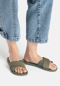 Grüne Rutschsandalen mit einem geflochtenen Riemen, flacher Gummisohle und offenem Design, getragen mit hochgekrempelten hellblauen Jeans.