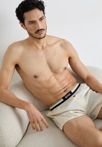 Lichtgekleurde boxershorts met een zwart en gouden gestreepte tailleband met het label "BOSS." Model heeft kort, donker haar en een gespierde bouw.