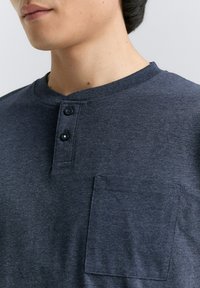 Námornícky modré henley tričko z jemného materiálu, s okrúhlym výstrihom, dvoma gombíkmi a ľavým vreckom na hrudi.