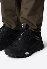 Baskets de course noires avec surfaces texturées, accents de couture jaunes et semelle en caoutchouc noire. Logo sur le talon, design à lacets, matériau GORE-TEX.