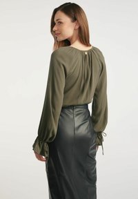 Oliwkowa bluzka z długimi rękawami i marszczonym tyłem, zestawiona z dopasowaną czarną skórzaną spódnicą. Bluzka ma wiązania na mankietach.