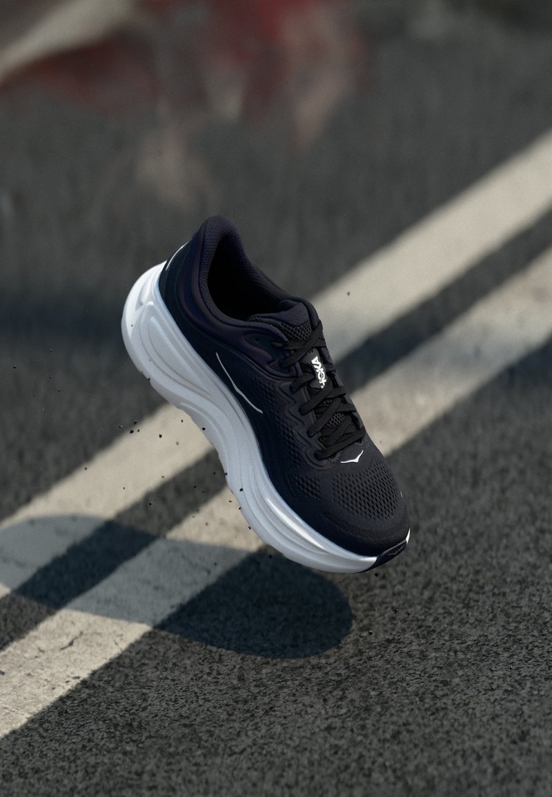 Baskets de sport noires avec une tige en maille, semelle blanche rembourrée, surface texturée et lacets noirs, positionnées au-dessus d'une route texturée.