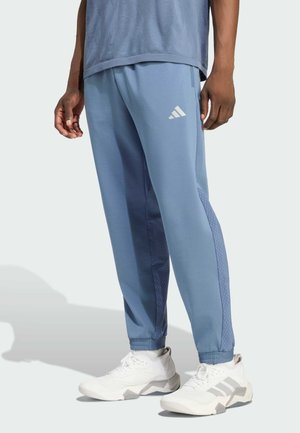 Man met lichtblauwe Adidas trainingsbroek en witte Adidas sneakers, staand tegen een effen lichte achtergrond.