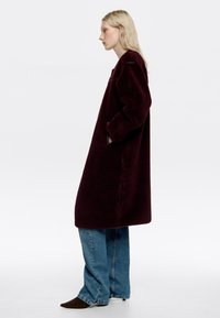 Abrigo burdeos de gran tamaño hecho de tela texturizada, con mangas largas y bolsillos laterales, combinado con jeans azules sueltos y zapatos marrones de puntera.