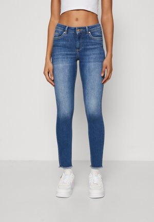 Mujer vestida con jeans skinny de tiro medio, de un azul desvaído y con dobladillos deshilachados, camiseta corta blanca, calcetines blancos y zapatillas blancas, de pie sobre un suelo gris.