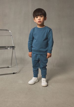 Noor poiss sinises dressipluusis ja pükstes valgete tossudega seisab lihtsa halli tausta ees metalltooli juures.