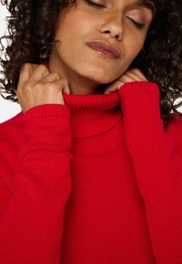 Pull en maille rouge avec col roulé, présentant une texture côtelée, doté de longues manches et d'un col haut, mis en valeur par une vue en gros plan.