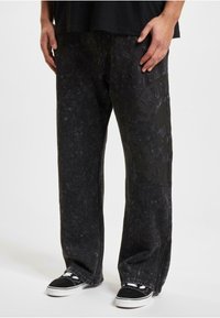 Sorte denim bukser med vid ben og teksturert overflate, med et stort svart grafisk trykk på det ene benet og glidelåser ved anklene.