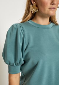 Miętowozielony, krótki top z bufkami na rękawach i okrągłym dekoltem. Miękka, gładka faktura; ozdobiony dużymi, dekoracyjnymi kolczykami w kształcie kwiatów.