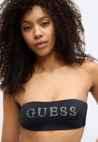 Μαύρο strapless μπικίνι τοπ με ρυτίδες· διακοσμημένο με στρας που σχηματίζουν τη λέξη "GUESS" με έντονη, καθαρή γραμματοσειρά. Ομαλή υφή, ελαστικό υλικό.