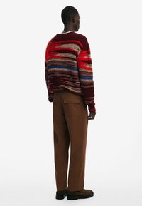 Sweter dziergany w czerwone, bordowe i niebieskie paski, w zestawieniu z brązowymi spodniami o szerokich nogawkach i ciemnymi butami slip-on. Tekstura wydaje się miękka, design jest swobodny.