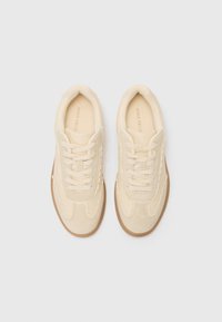 Beige suede sneakers with a textured surface, round toe, and contrasting brown rubber outsole. Features laces and decorative stitching.