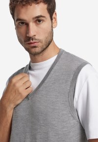 Grå stickad ärmlös tröja med V-ringning, lager-på-lager över en vit T-shirt. Mjuk textur, figursydd design, minimala sömnadsdetaljer.