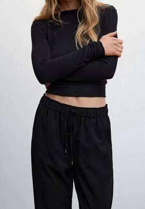 Vrouw met een zwart cropped shirt met lange mouwen en zwarte broek met koord, armen gekruist, licht golvend haar over de schouders, neutrale achtergrond.