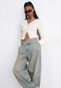 Bjela rebrasta dugorukava crop majica s prednjim zatvaračem, uparena s svijetloplavim oversized trapericama. Model nosi tamne sunčane naočale.
