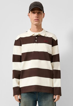 Jonge man met een zwarte pet en een poloshirt met lange mouwen met brede bruine en witte horizontale strepen, staand tegen een effen achtergrond.